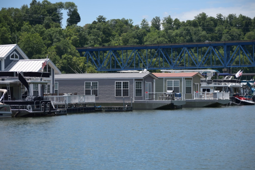 A row of houseboats along a calm lake, with a blue bridge and green trees in the background.