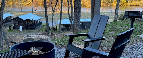 A cozy outdoor scene featuring a fire pit, wooden chairs, and cabins by a river surrounded by trees.