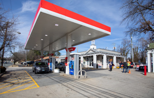 A gas station with a red canopy, cars at the pumps, and people in safety vests near the entrance on a clear day.