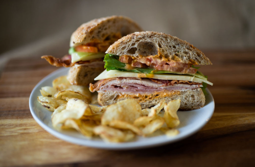 A sandwich with layers of bacon, lettuce, tomato, and ham on whole grain bread, served with a side of potato chips.