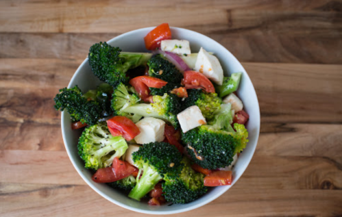 A bowl of fresh broccoli, tomatoes, and diced cheese on a wooden surface.