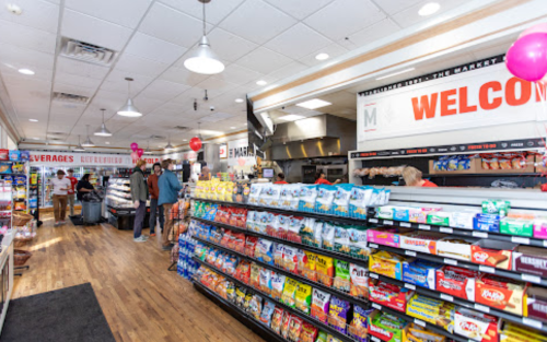 Interior of a convenience store with shelves stocked with snacks, drinks, and a welcoming sign. Customers are browsing.
