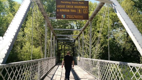 A person walks on a metal bridge with a sign for Locust Creek Boardwalk and Riparian Trail in a lush green park.