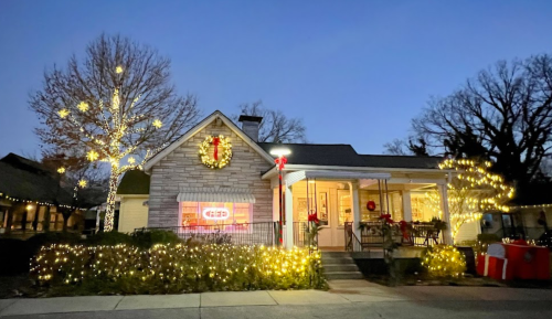 A cozy house decorated with festive lights and a wreath, surrounded by illuminated trees, at dusk.