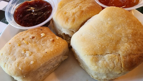 Three fluffy biscuits served with two small cups of fruit preserves on a white plate.