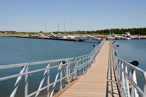 A wooden dock extends over calm water, leading to a marina filled with boats and surrounded by trees under a clear sky.