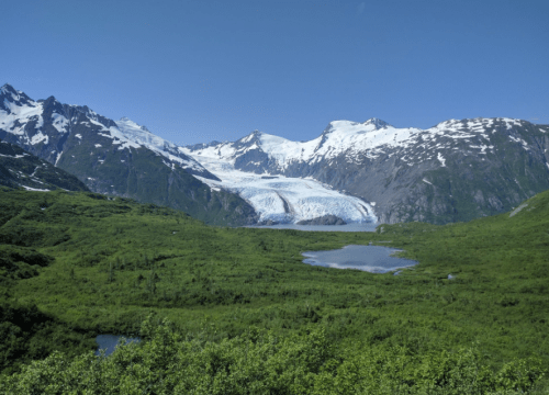 A stunning landscape featuring a glacier surrounded by snow-capped mountains and lush green valleys under a clear blue sky.