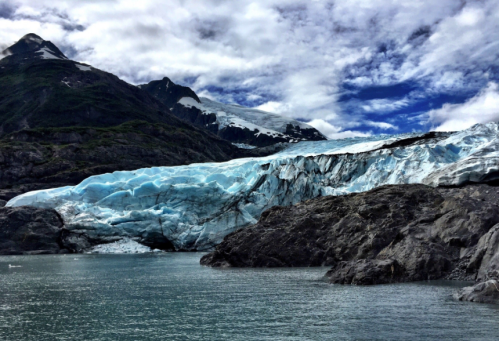 A glacier with blue ice flows into a calm body of water, surrounded by rocky terrain and mountains under a cloudy sky.