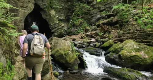 A hiker approaches a cave entrance beside a flowing stream and lush greenery.