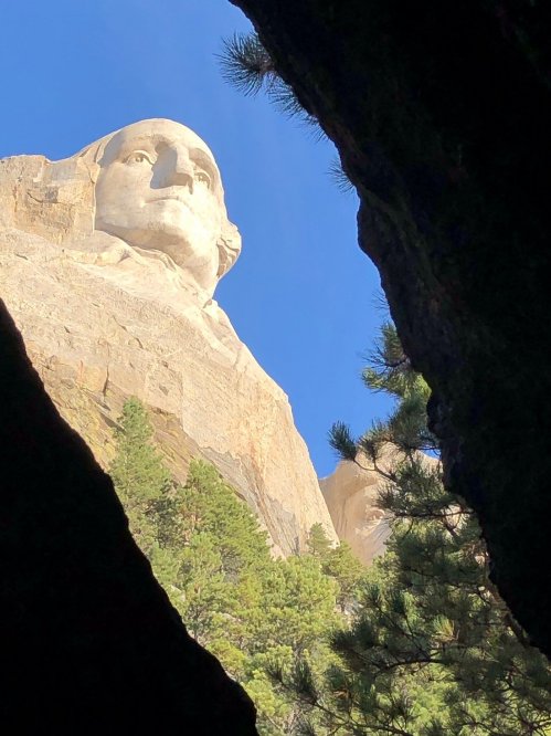 View of Mount Rushmore's George Washington from a rocky crevice, framed by trees and a clear blue sky.