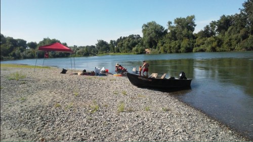 A sandy riverbank with a tent, people relaxing, and a canoe by the water surrounded by lush greenery.