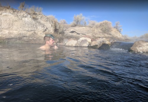 A person relaxes in a steaming hot spring surrounded by rocky terrain and sparse vegetation under a clear blue sky.