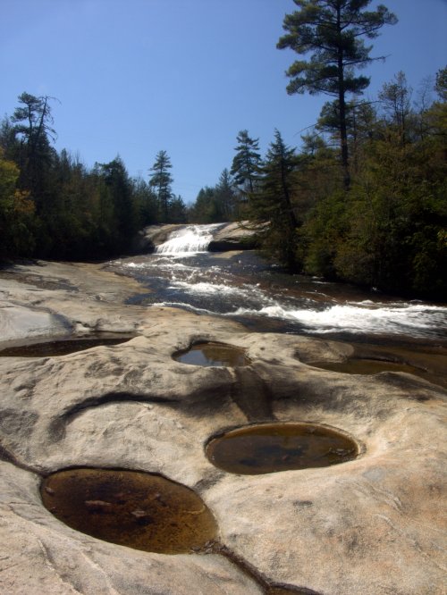 A serene river flows over smooth rocks, with small pools and trees lining the banks under a clear blue sky.