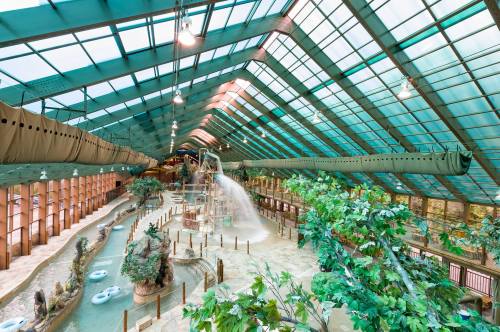 Indoor water park with a glass roof, featuring slides, a lazy river, and lush greenery throughout the space.