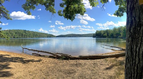 A serene lake surrounded by trees, with a sandy shore and blue sky dotted with clouds.