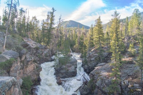 A rushing river flows through rocky terrain, surrounded by lush green trees and mountains under a blue sky.