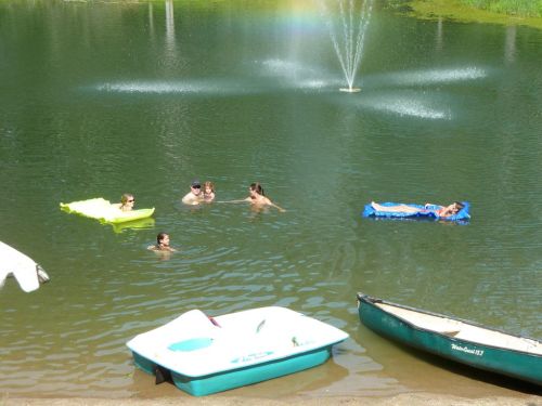People swimming and relaxing on floats in a pond, with a fountain in the background and boats nearby.