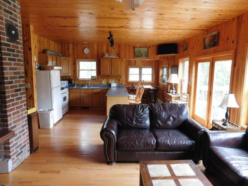 Cozy wooden cabin interior featuring a living area with a leather sofa, kitchen, dining space, and large windows.