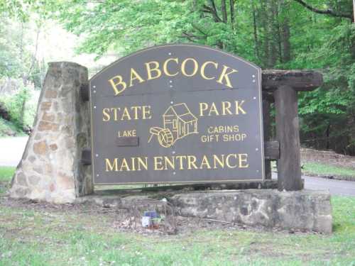 Sign for Babcock State Park, featuring "Main Entrance" and details about cabins and a gift shop, surrounded by trees.