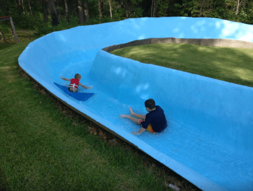 Two children play in a blue water slide, one sliding down while the other sits in the shallow water at the bottom.