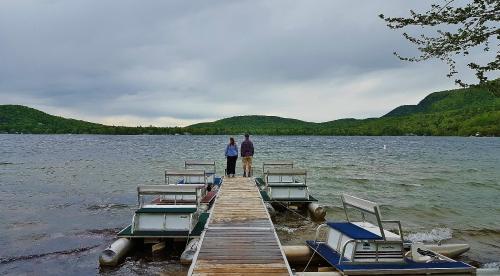 Two people stand on a wooden dock overlooking a calm lake surrounded by green hills under a cloudy sky.
