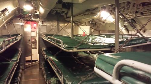 Interior of a submarine showing metal bunk beds with green mattresses, dim lighting, and metal walls.