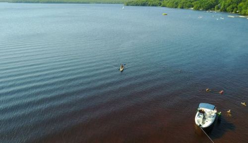 Aerial view of a calm lake with a kayak and a boat, surrounded by greenery and people swimming nearby.