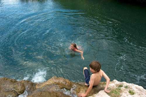 Two children play in a natural swimming area, one swimming in the water and the other sitting on a rock by the edge.