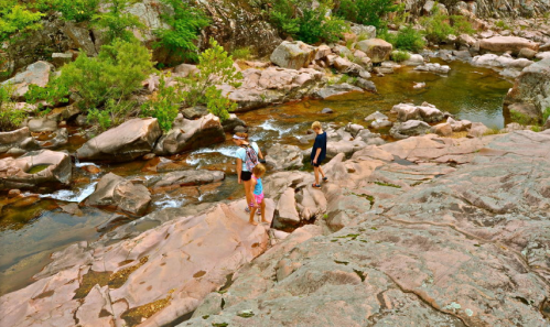 Three people walking along rocky terrain by a shallow stream, surrounded by greenery and natural rock formations.