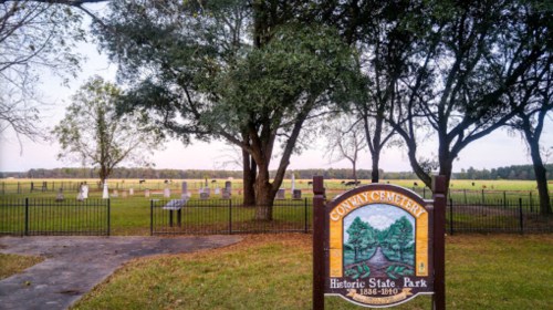 Sign for Conway Cemetery at Historic State Park, surrounded by trees and gravestones in a grassy field.