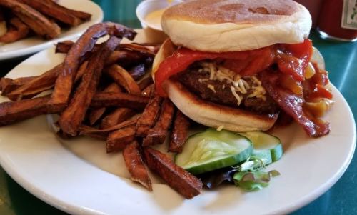 A plate with a burger topped with bacon and sauce, served with crispy sweet potato fries and cucumber slices.