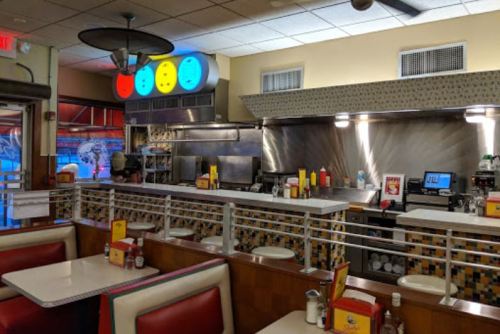 A retro diner interior featuring red and white booths, a colorful light fixture, and a visible kitchen area.