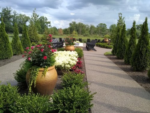 A landscaped pathway lined with potted flowers and shrubs, leading to seating areas among greenery and trees.
