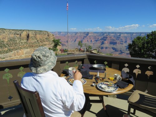 A person in a white robe and hat sits at a table with breakfast, overlooking the Grand Canyon under a clear blue sky.