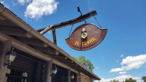 A wooden sign reading "El Tovar" hangs from a rustic building under a blue sky with scattered clouds.