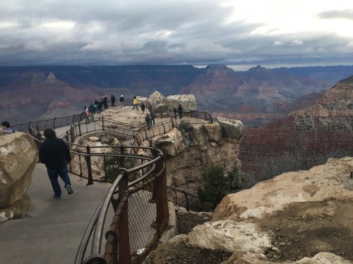 Visitors walk along a rocky overlook at the Grand Canyon, with dramatic cliffs and cloudy skies in the background.