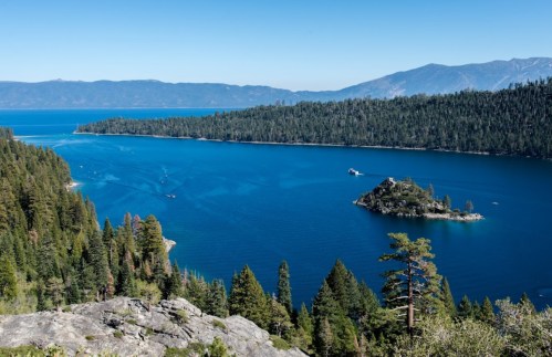 A scenic view of a blue lake surrounded by lush green forests and mountains under a clear blue sky.
