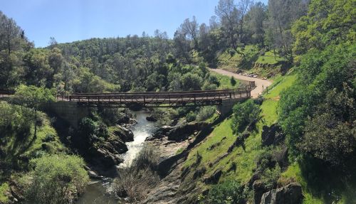 A wooden bridge spans a river surrounded by lush greenery and hills under a clear blue sky.