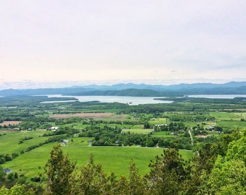 A panoramic view of lush green fields and a lake surrounded by mountains under a cloudy sky.