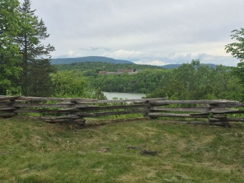 A wooden fence in the foreground overlooks a lush green landscape and a lake, with mountains in the background.