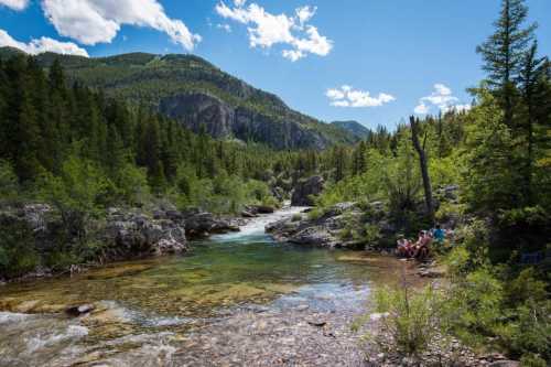 A serene river flows through a lush green landscape, with mountains in the background and people sitting by the water.