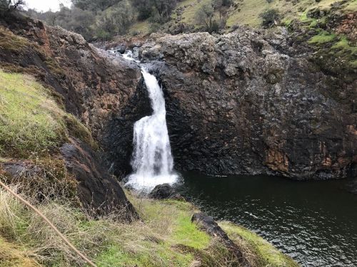 A waterfall cascading over rocky cliffs into a serene pool, surrounded by lush greenery and rugged terrain.