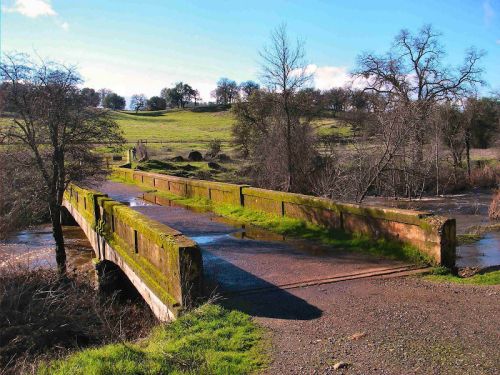 A moss-covered bridge spans a small stream, surrounded by green fields and trees under a clear blue sky.