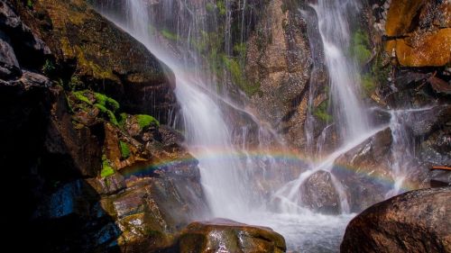 A vibrant rainbow arcs over a cascading waterfall surrounded by moss-covered rocks.