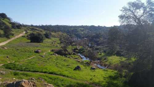 A scenic landscape featuring green hills, a winding stream, and rocky outcrops under a clear blue sky.