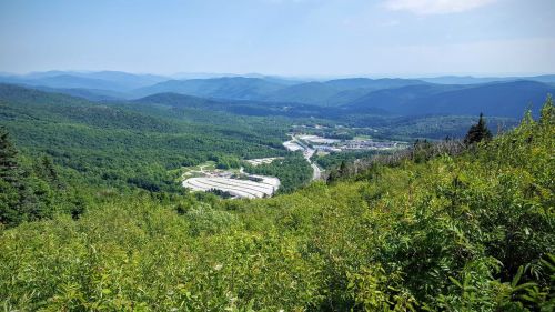 A panoramic view of lush green mountains and valleys under a clear blue sky, with a winding road visible below.