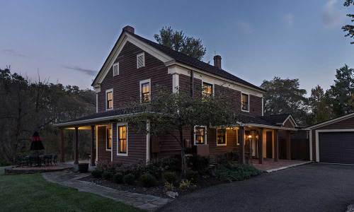 A charming two-story house with a dark exterior, illuminated windows, and a cozy porch, surrounded by trees at dusk.