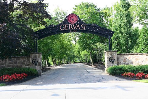 Entrance archway to Gervasi Vineyard, surrounded by trees and colorful flowers, with a paved driveway leading in.