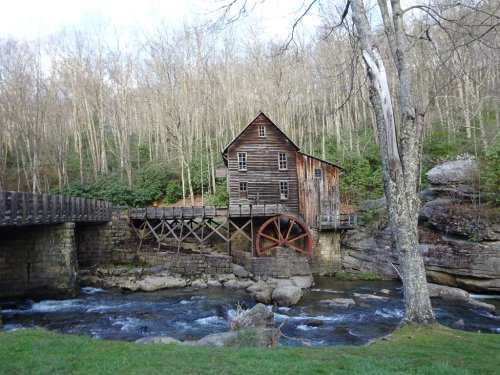 A rustic wooden mill with a water wheel beside a flowing stream, surrounded by trees and rocky terrain.
