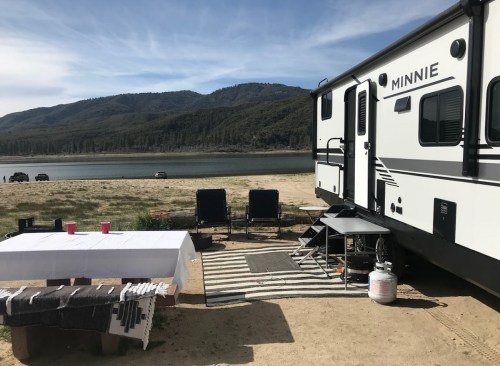 A camper by a lake with mountains in the background, featuring outdoor seating and a table set up nearby.